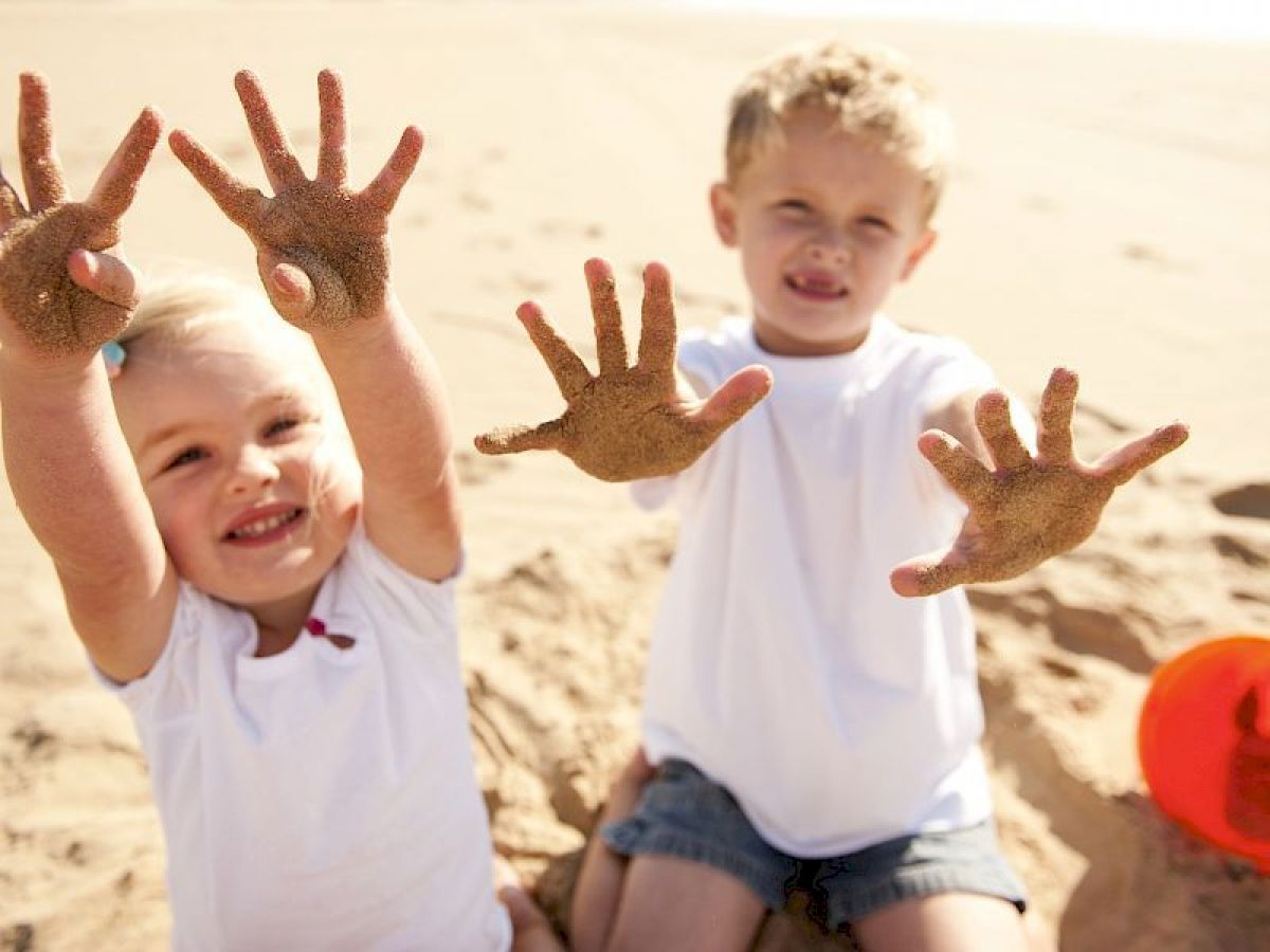 Two smiling children at the beach, holding up their sandy hands. They are sitting in the sand with a red bucket nearby.