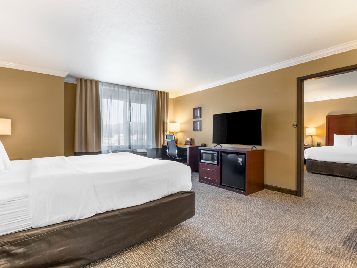 This image shows a hotel room with a king-sized bed, a TV, a desk, a microwave, a window, and an adjoining room with another bed.