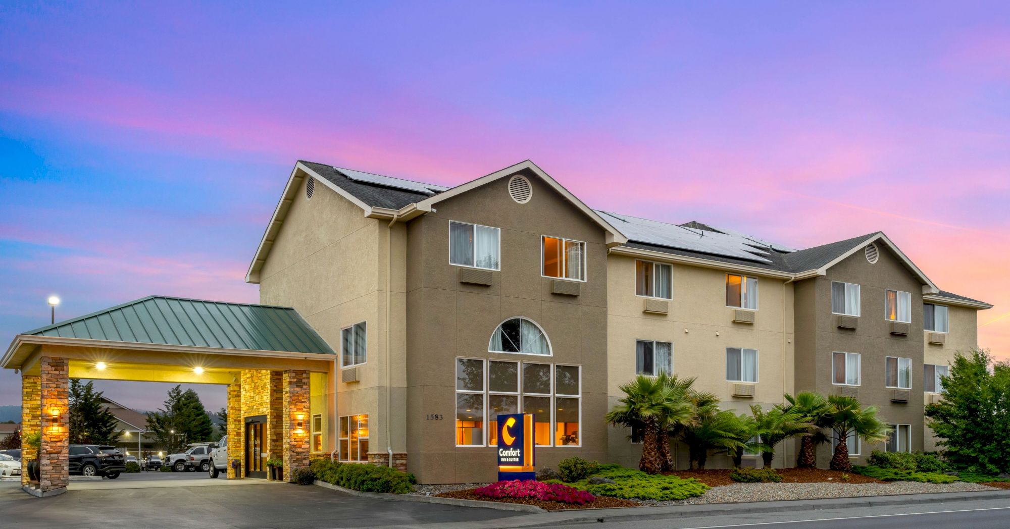 The image shows a three-story hotel building with the Comfort Inn logo, a portico, landscaped garden, and a vibrant sunset sky in the background.