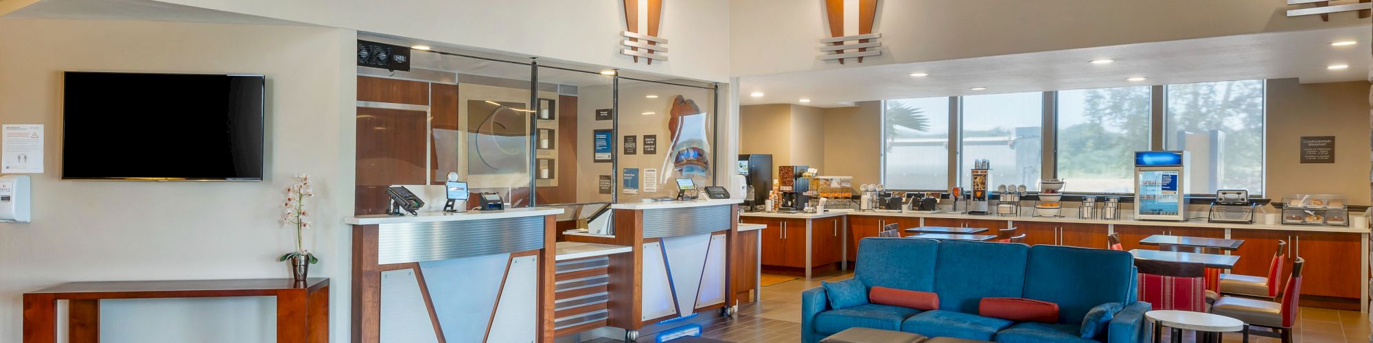 The image shows a modern lobby area with a reception desk, seating, a TV, and a breakfast bar. The space features contemporary decor and ample lighting.