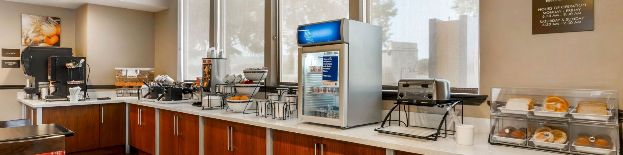 The image shows a hotel breakfast area with a coffee machine, juice dispenser, toaster, and various food items on the countertop.