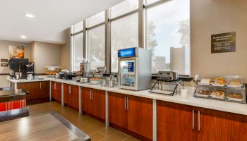 A breakfast buffet area with coffee machines, a toaster, an ice machine, and a variety of food items including bagels and cereals on the counter.