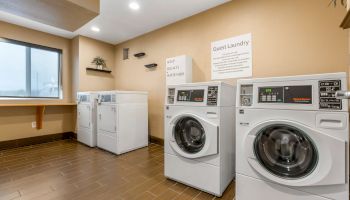 The image shows a clean and organized laundry room with washing machines and dryers, a folding table, and signs on the wall.