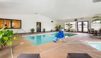The image shows an indoor swimming pool area with poolside chairs, a hot tub, plants, and a hoist for disabled access, in a bright, clean space.