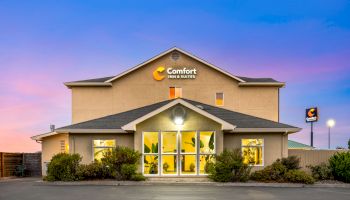 The image shows the exterior of a Comfort Inn & Suites building with a lit-up logo, large front windows, a parking area, and some bushes.