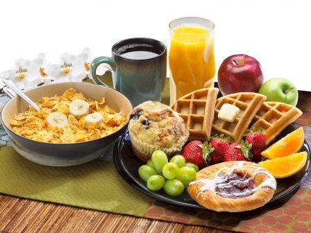 A hearty breakfast spread featuring cornflakes with bananas, coffee, orange juice, apples, waffles with butter, grapes, strawberries, muffin, and pastry.