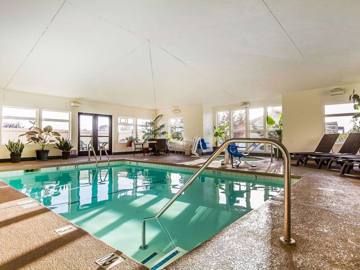 An indoor pool area with lounge chairs, potted plants, large windows, and a handrail leading into the water.